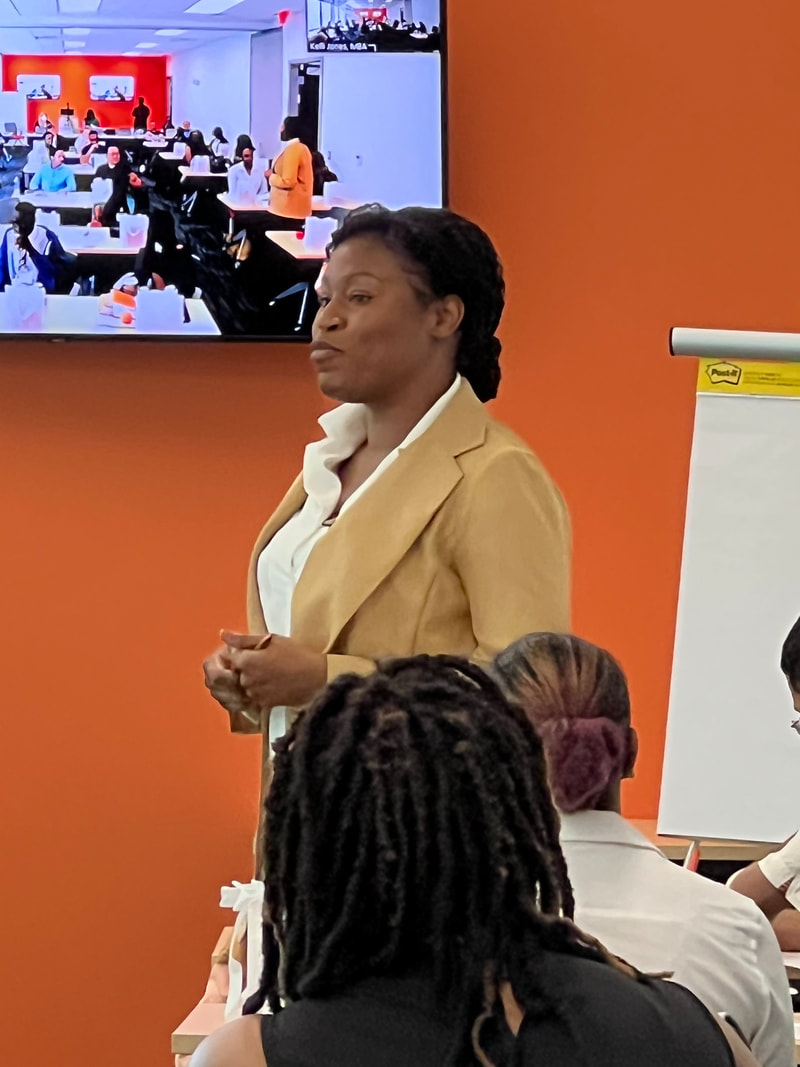 Participant in a mustard jacket addressing the group in front of a wall-mounted screen