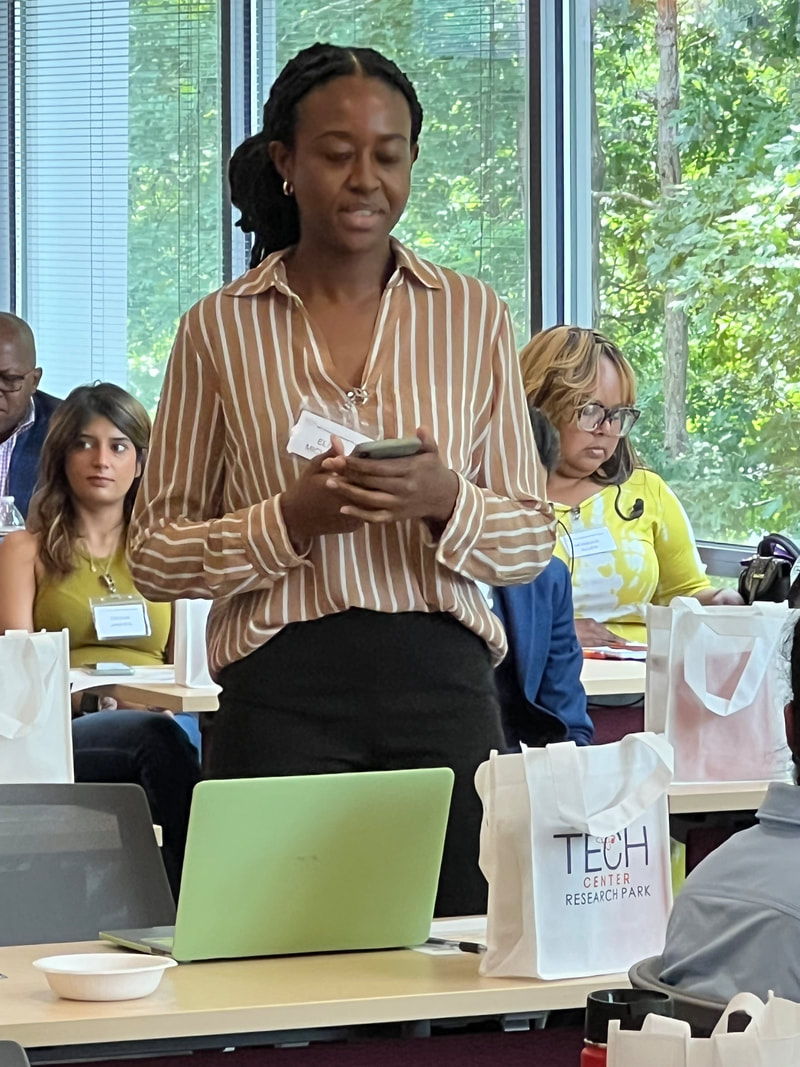 Participant in a striped blouse addressing workshop attendees beside the windows