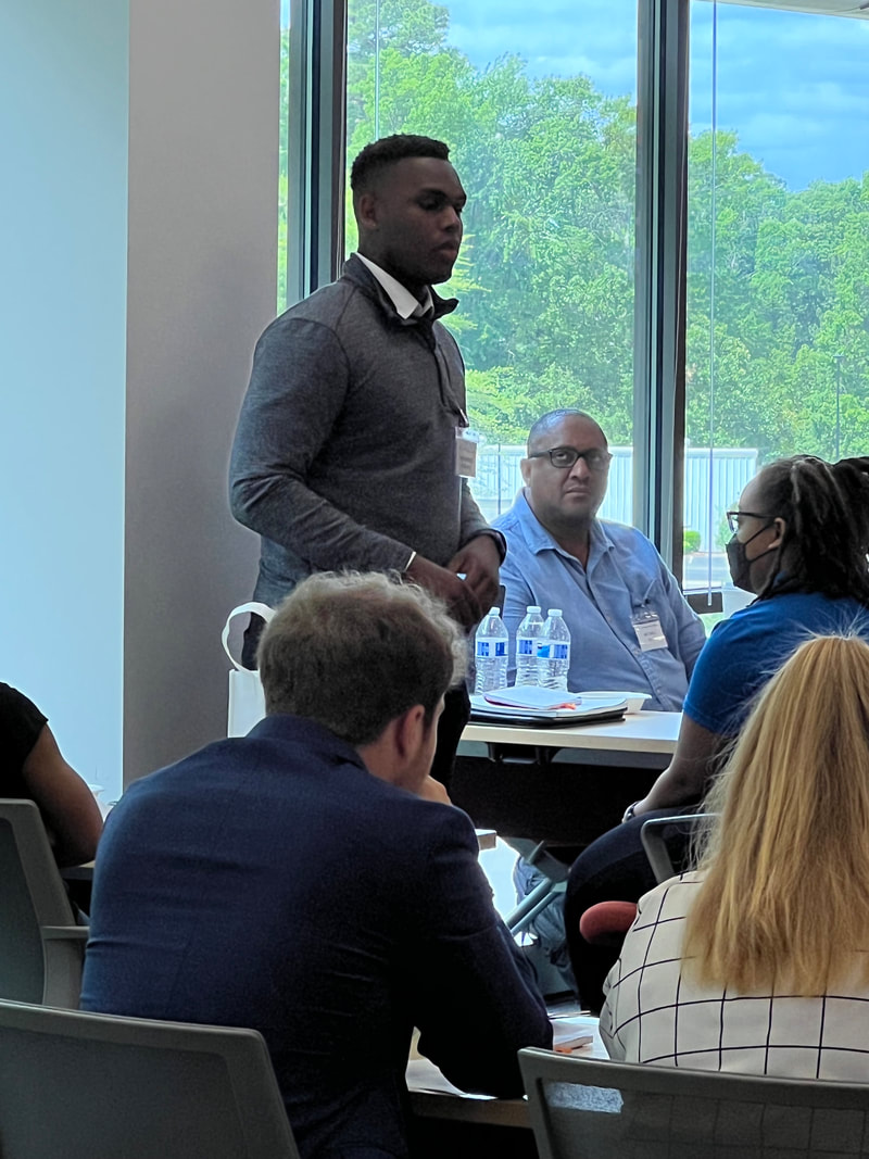 Participant in a gray sweater speaking beside a table of seated attendees