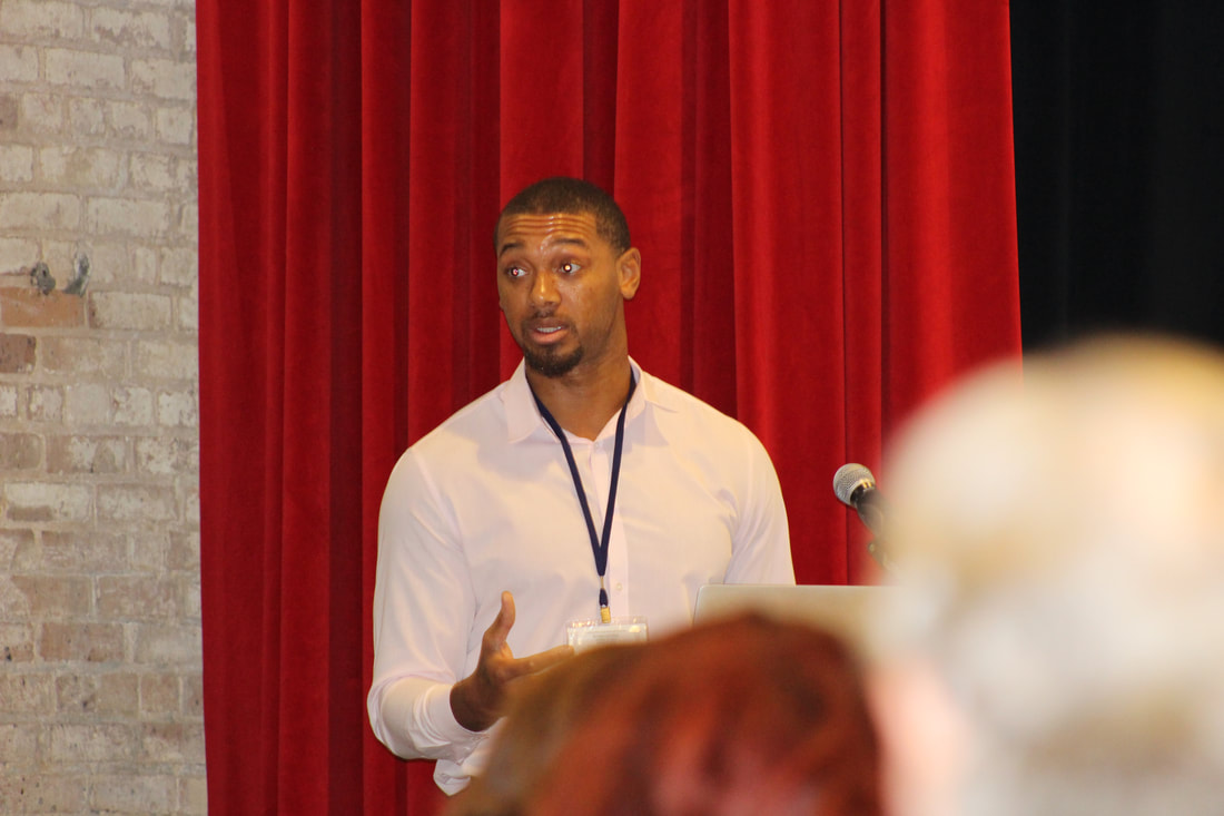 Participant in a white shirt speaking into a microphone before red stage curtains