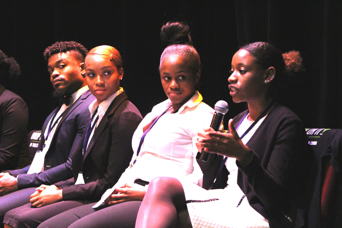 Four students seated on stage during an interview, with one speaking into a microphone